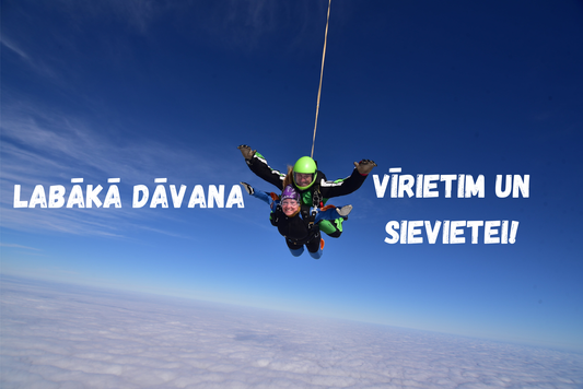 Tandēma izpletņlēciens virs mākoņiem ar skaidrām debesīm. Instruktors un pasažiere smaida brīvajā kritienā. Teksts attēlā: Labākā dāvana vīrietim un sievietei.